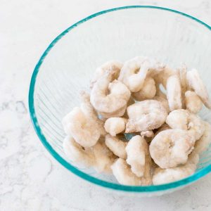 A clear mixing bowl is filled with frozen raw shrimp.