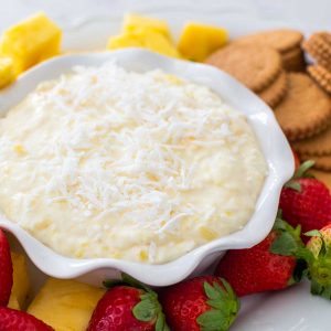 A ruffled bowl of pina colada dip has shredded coconut on top and is surrounded by fresh strawberries, pineapple, and gingersnap cookies.
