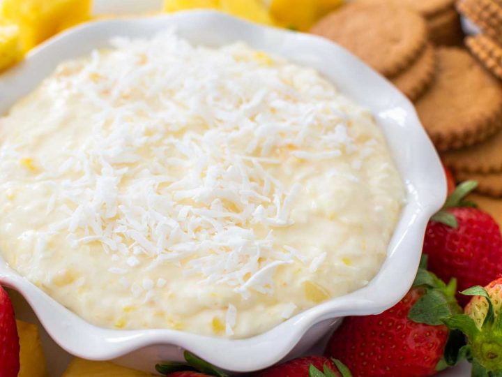 A ruffled bowl of pina colada dip has shredded coconut on top and is surrounded by fresh strawberries, pineapple, and gingersnap cookies.
