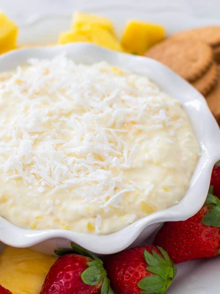A ruffled bowl of pina colada dip has shredded coconut on top and is surrounded by fresh strawberries, pineapple, and gingersnap cookies.