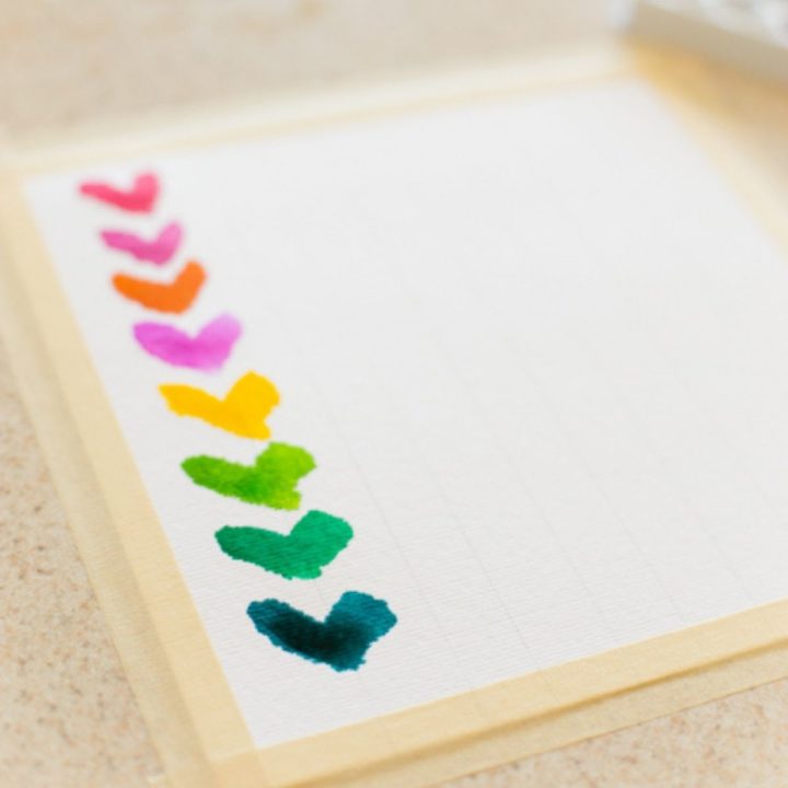 A row of rainbow colored watercolor hearts have been painted on white paper.
