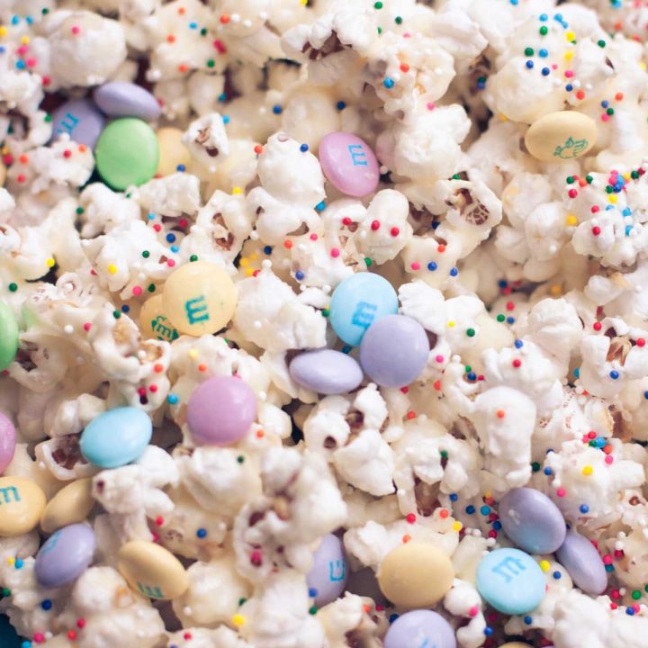 A batch of birthday cake popcorn has pastel M&Ms and rainbow sprinkles with a white candy coating.