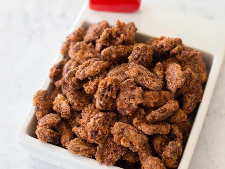 A dish of candied nuts has a red heart decoration. The nuts are coated in a thick sugar crust.