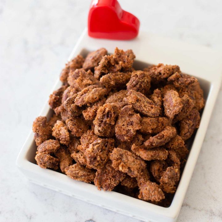 A dish of candied nuts has a red heart decoration. The nuts are coated in a thick sugar crust.