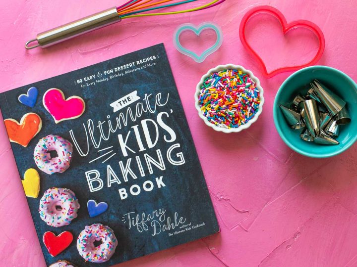 A copy of The Ultimate Kids' Baking Book sits on a pink background with a whisk, sprinkles, and cookie cutters.