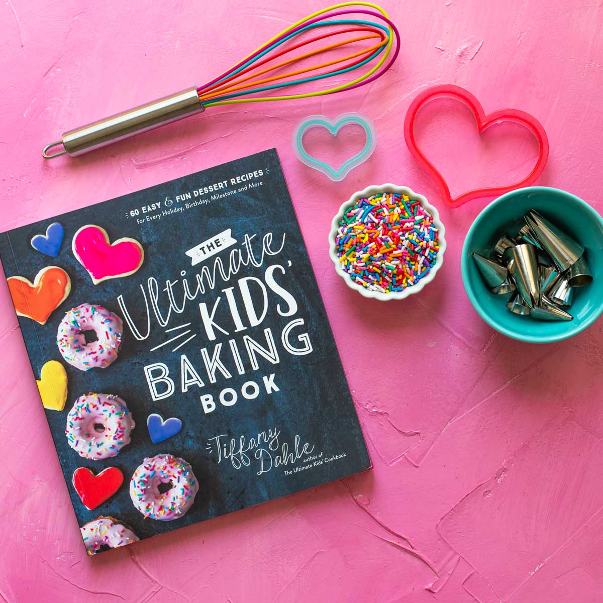 A copy of The Ultimate Kids' Baking Book sits on a pink background with a whisk, sprinkles, and cookie cutters.