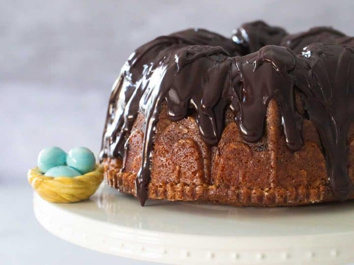 A chocolate glazed bundt cake sits on a white cake plate with a blue robins egg decoration to the left.
