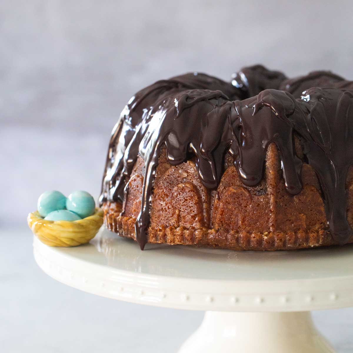 A chocolate glazed bundt cake sits on a white cake plate with a blue robins egg decoration to the left.