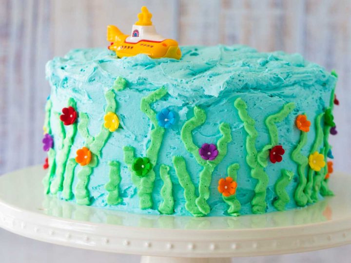 A light blue birthday cake has been decorated to look like an under the sea garden and has a yellow submarine on top.