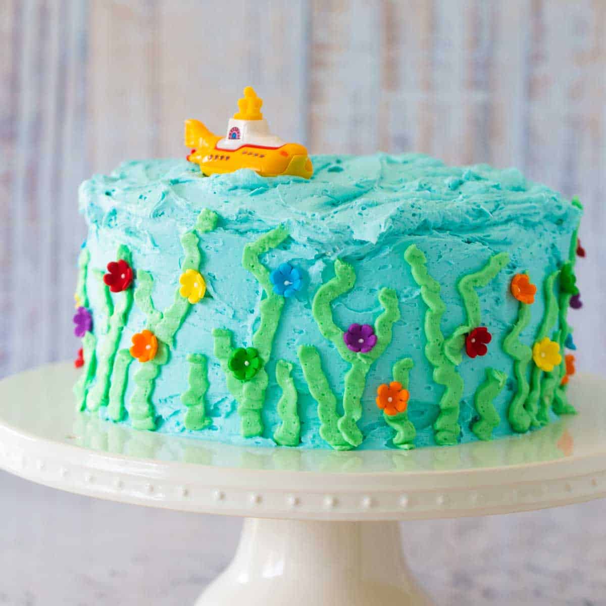 A light blue birthday cake has been decorated to look like an under the sea garden and has a yellow submarine on top.