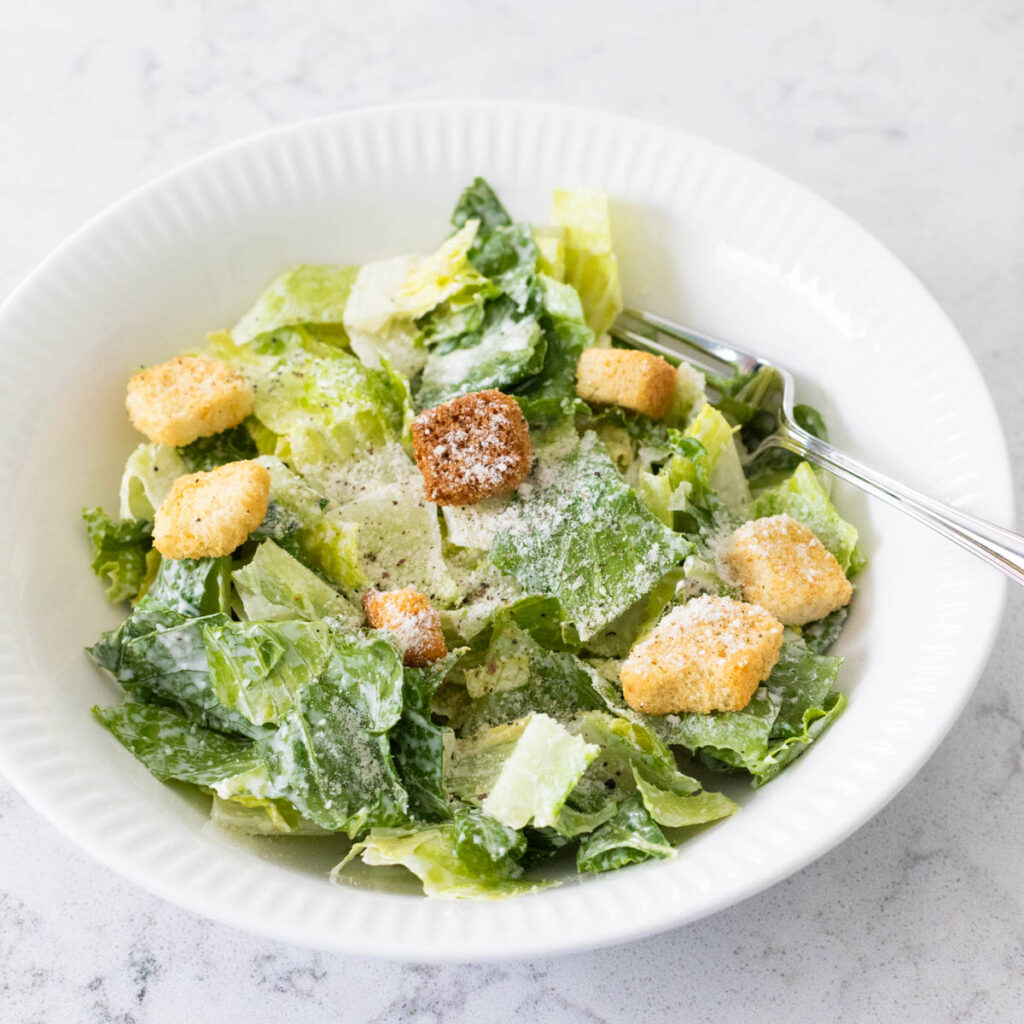 A dressed Caesar salad with croutons and grated parmesan is in a white bowl with a fork.