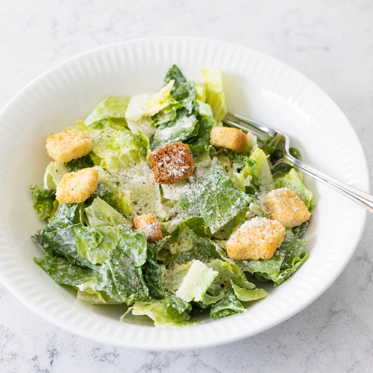 A dressed Caesar salad with croutons and grated parmesan is in a white bowl with a fork.
