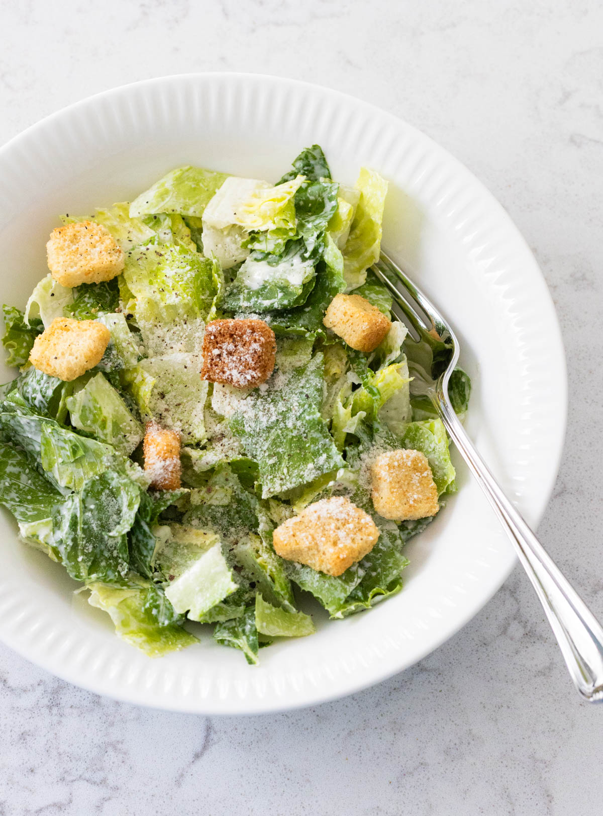 A dressed Caesar salad with croutons and grated parmesan is in a white bowl with a fork.
