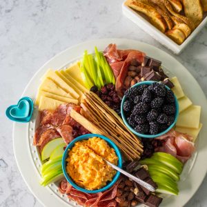A round snack board has sliced green apples, spreadable cheese, slices of cheese, ruffled slices of various meats, and a bowl of fresh blackberries.