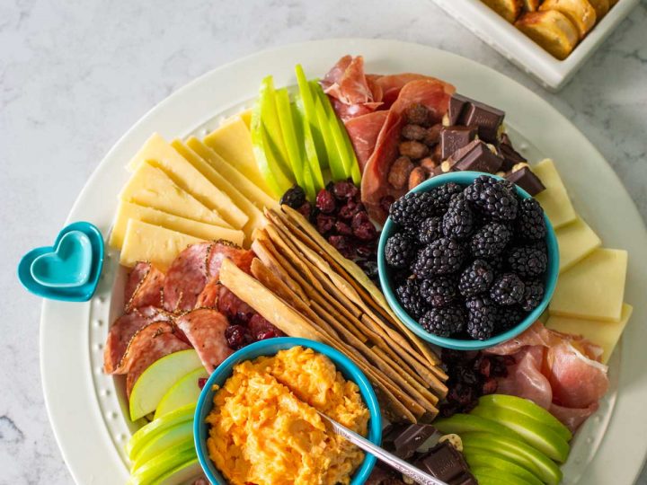 A round snack board has sliced green apples, spreadable cheese, slices of cheese, ruffled slices of various meats, and a bowl of fresh blackberries.