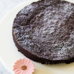 A rich chocolately round torte is on a cake plate with a pink ceramic flower accent. Powdered sugar is dusted over the top.