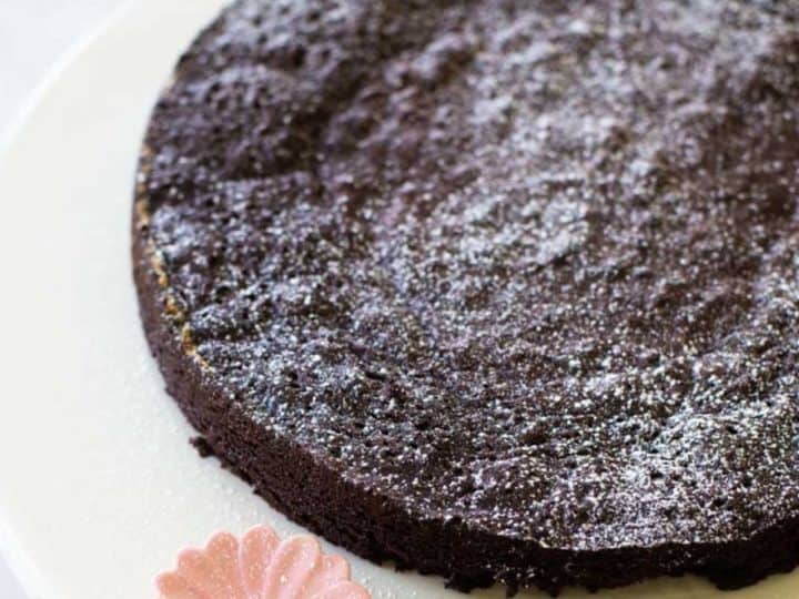 A rich chocolately round torte is on a cake plate with a pink ceramic flower accent. Powdered sugar is dusted over the top.