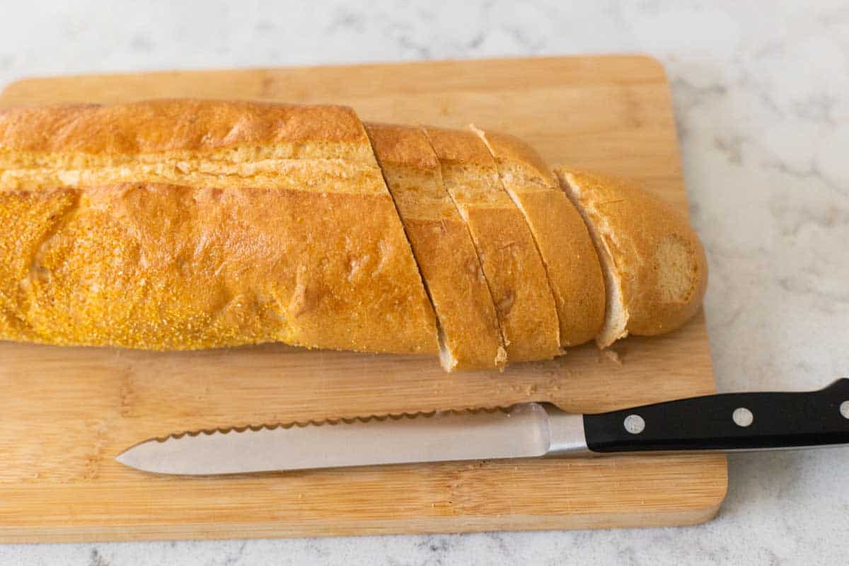 A serrated knife sits on the cutting board next to a loaf of bread that has been sliced on an angle. 