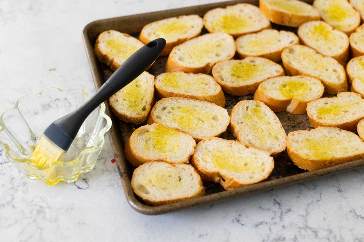 The bread slices are in an even layer on the baking sheet. A cup of olive oil has a pastry brush dipped into it.