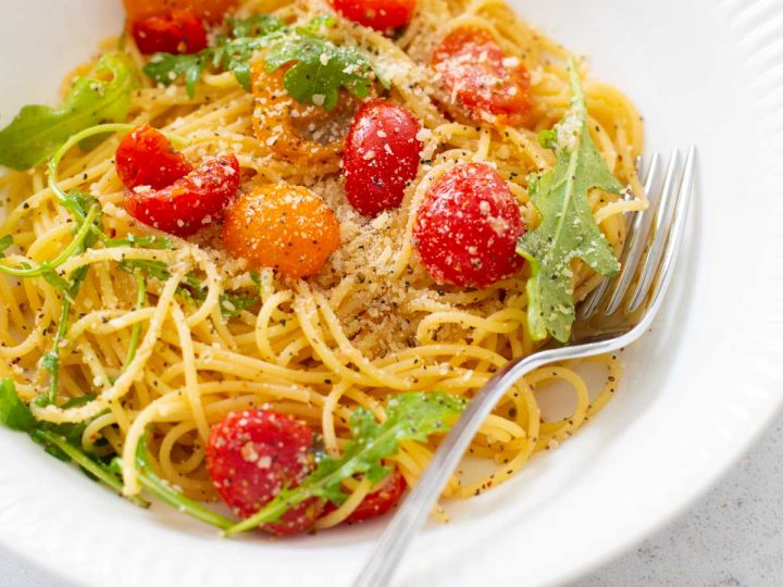 A white bowl is filled with thin spaghetti and fresh cherry tomatoes and arugula. Parmesan is sprinkled over the top.