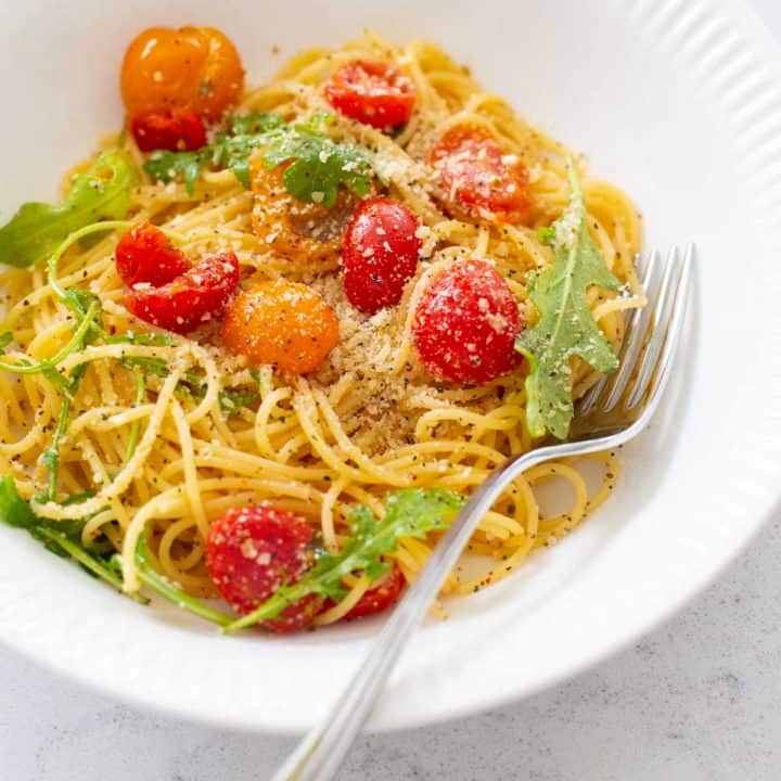 A white bowl is filled with thin spaghetti and fresh cherry tomatoes and arugula. Parmesan is sprinkled over the top.