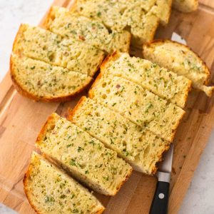 The baked garlic bread is on a cutting board and has been cut into wedges for serving.