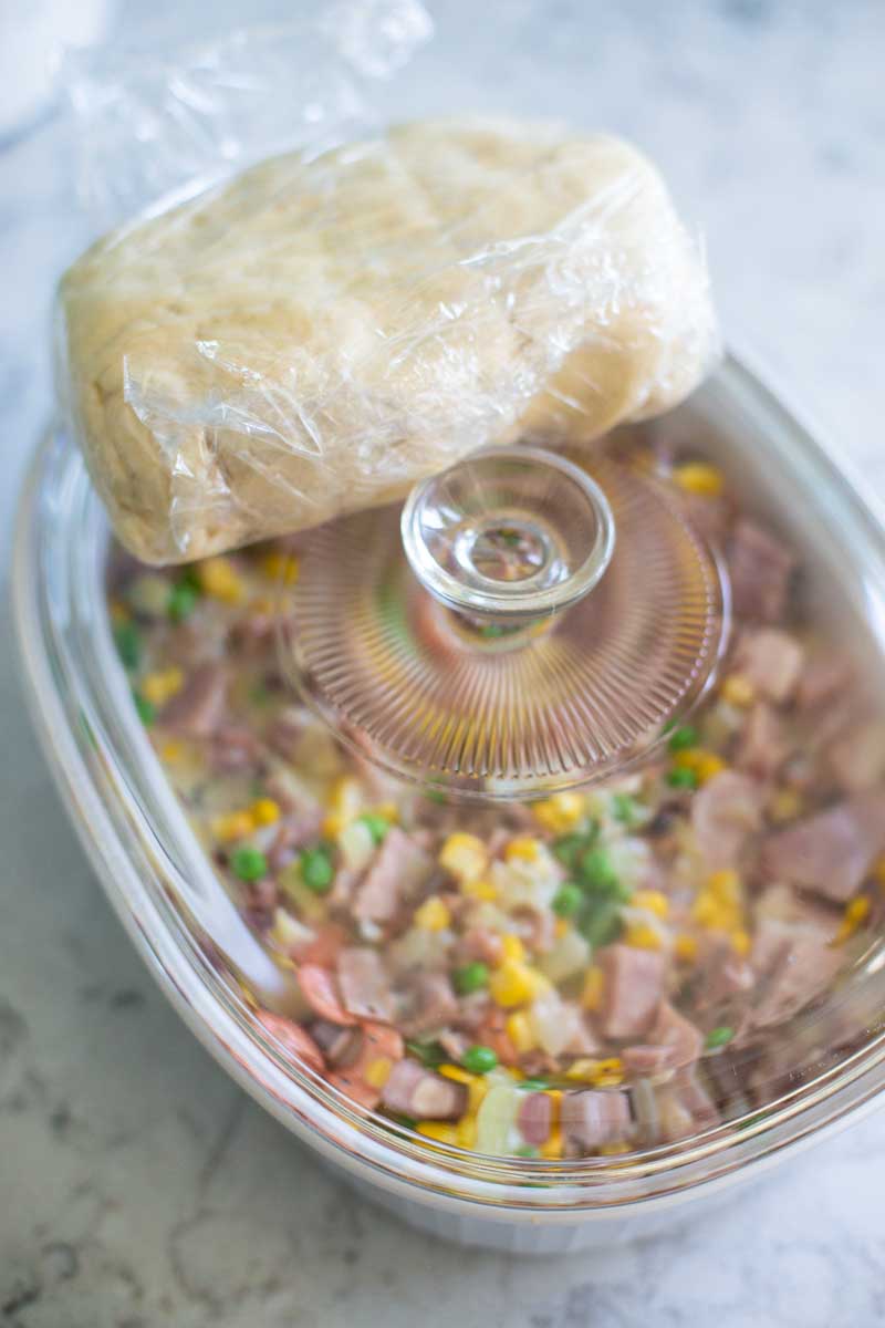 The pie crust dough is wrapped in plastic wrap and sitting on top of a storage container filled with the ham and veggie filling.