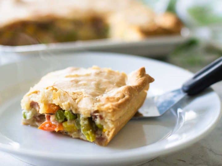 A slice of ham slab pot pie sits on a plate with a serving spatula. Big chunks of fresh veggies peek out from under a flaky pie crust.