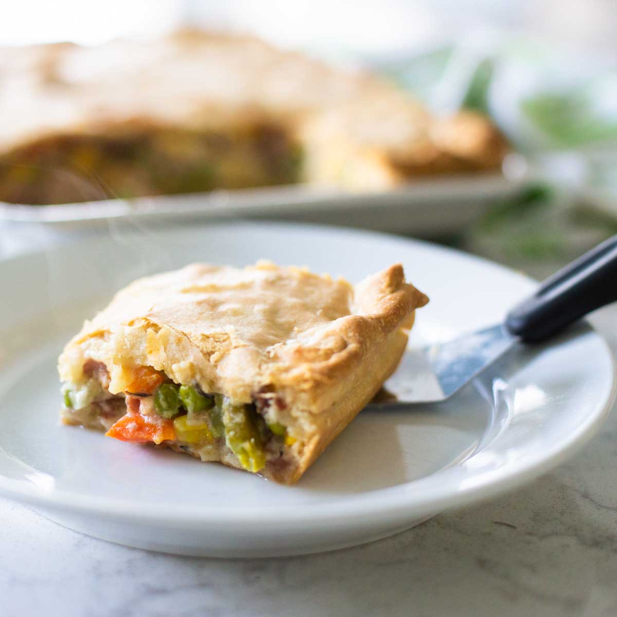 A slice of ham slab pot pie sits on a plate with a serving spatula. Big chunks of fresh veggies peek out from under a flaky pie crust.