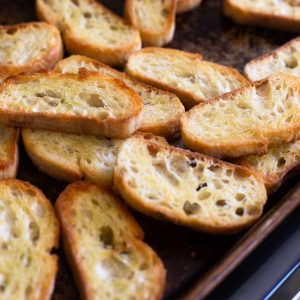 Homemade toasted crostini slices sit on a cookie sheet fresh from the oven. They are golden brown.