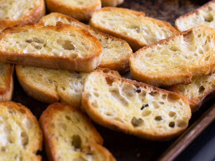 Homemade toasted crostini slices sit on a cookie sheet fresh from the oven. They are golden brown.