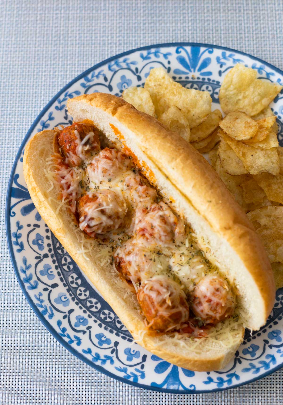 The baked meatball sub is on a blue plate next to some potato chips.