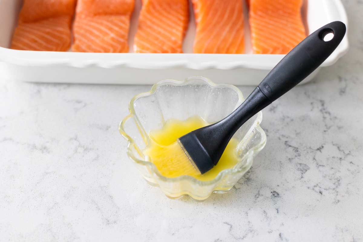 The bowl of melted butter has a pastry brush in it. The salmon is in the background.