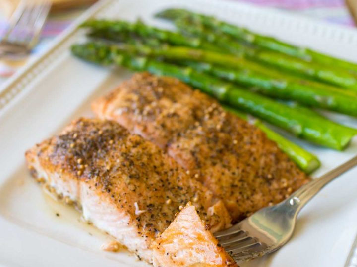 A platter of oven baked salmon next to roasted asparagus has a fork showing the flaky pink fish.