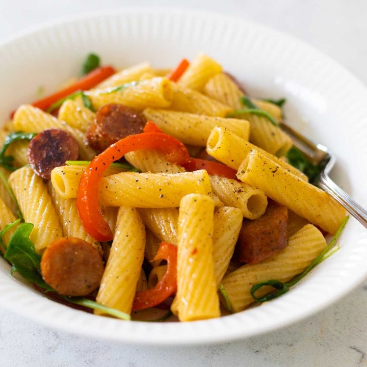 A white bowl has rigatoni pasta with sausage and peppers.