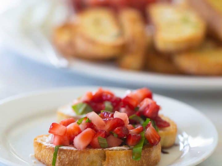 2 crostini toasts are topped with diced strawberries and a sprinkle of fresh basil to show the strawberry bruschetta appetizer. A plate of crostini sits in the background.