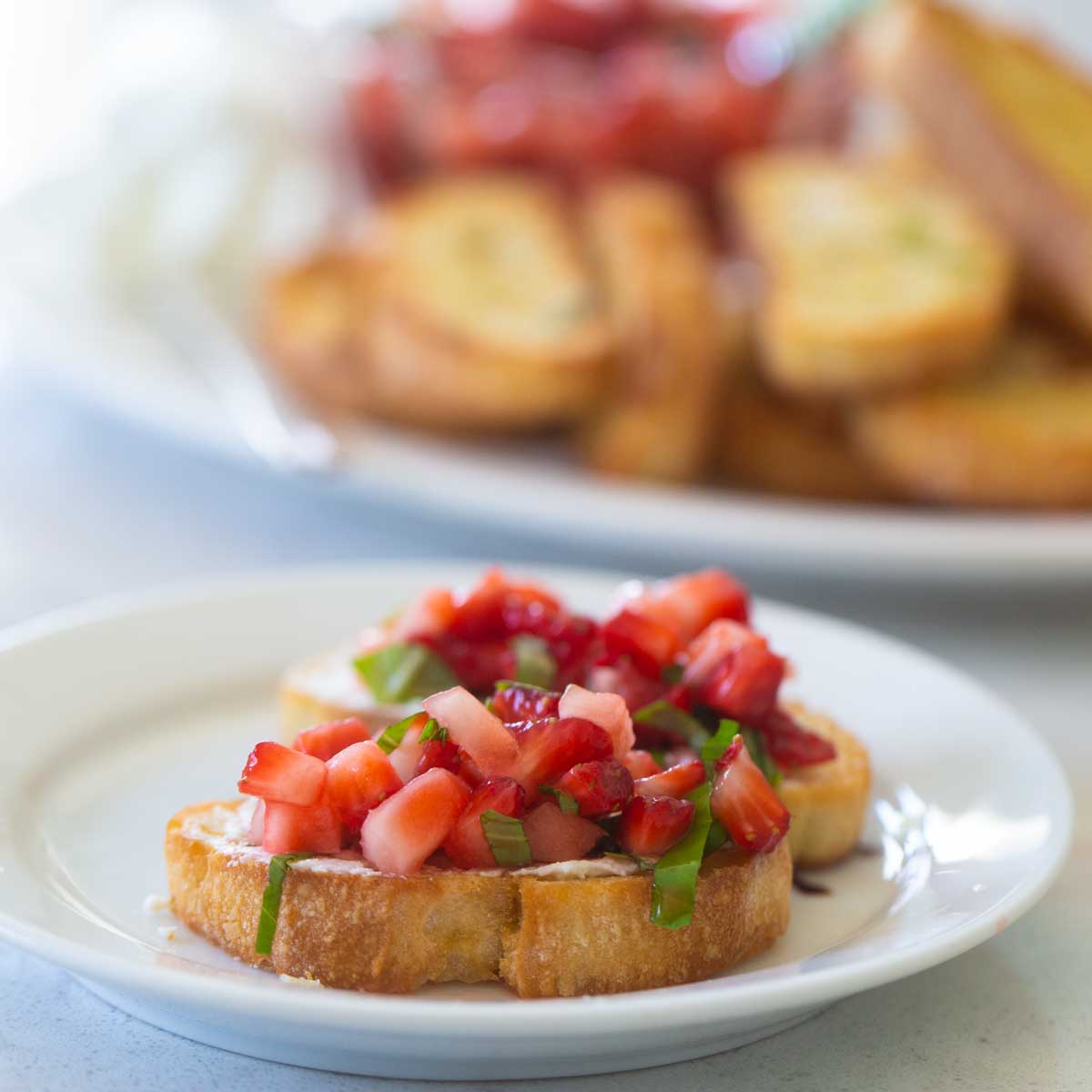2 crostini toasts are topped with diced strawberries and a sprinkle of fresh basil to show the strawberry bruschetta appetizer. A plate of crostini sits in the background.