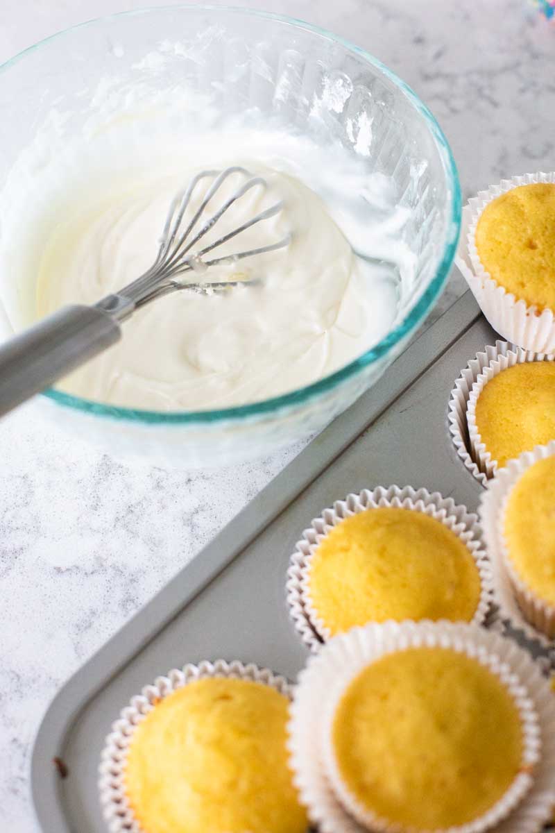 A bowl of vanilla glaze has a whisk and is sitting next to a muffin pan filled with vanilla cupcakes.