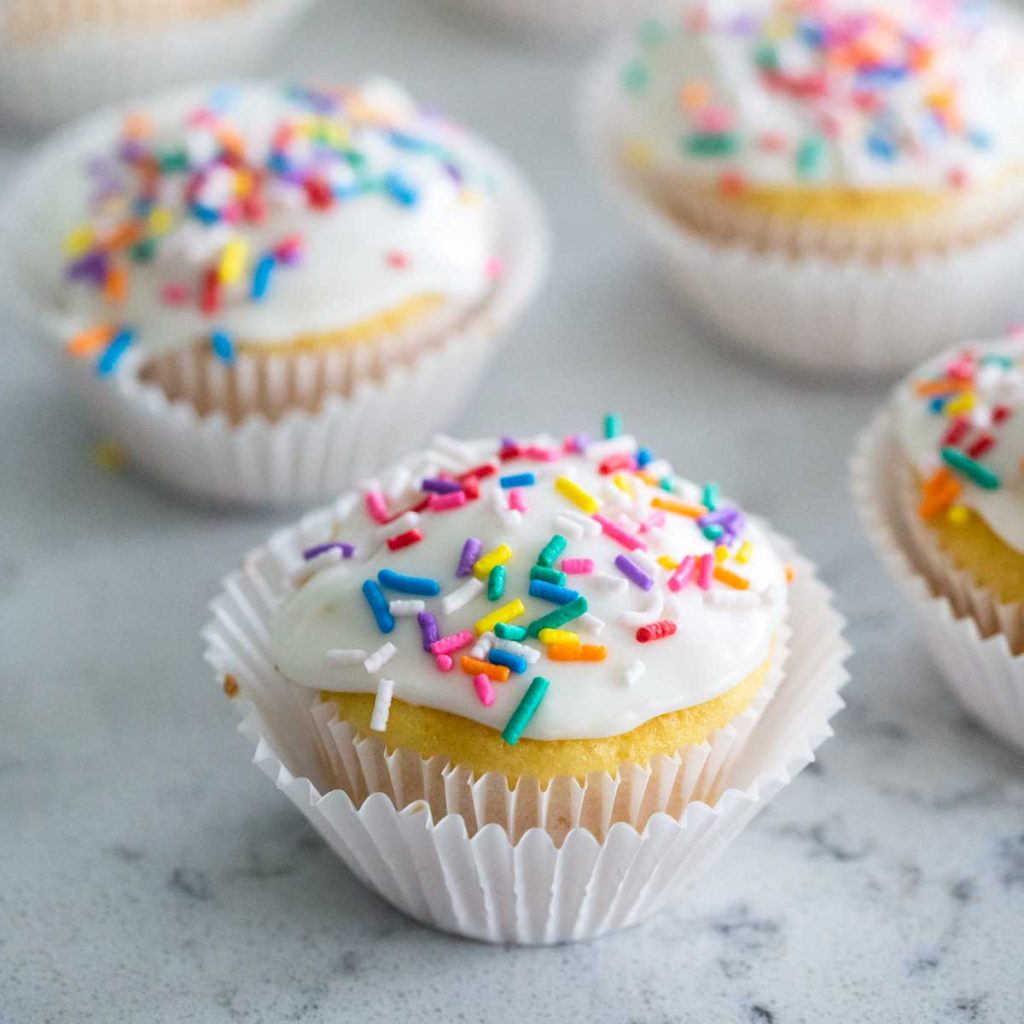 A vanilla cupcake in a white wrapper has white vanilla glaze and rainbow sprinkles on top. More cupcakes are in the background.