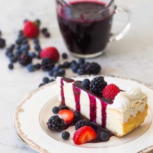 A slice of plain cheesecake has fresh strawberries, blackberries, and blueberries sprinkled over the top and on the table. A drizzle of dark purple triple berry sauce is dripping down from the top. A pitcher of sauce is nearby.