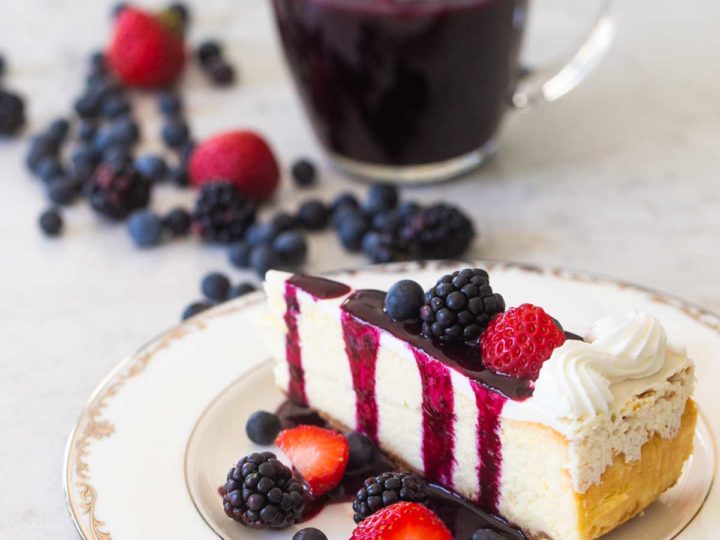 A slice of plain cheesecake has fresh strawberries, blackberries, and blueberries sprinkled over the top and on the table. A drizzle of dark purple triple berry sauce is dripping down from the top. A pitcher of sauce is nearby.