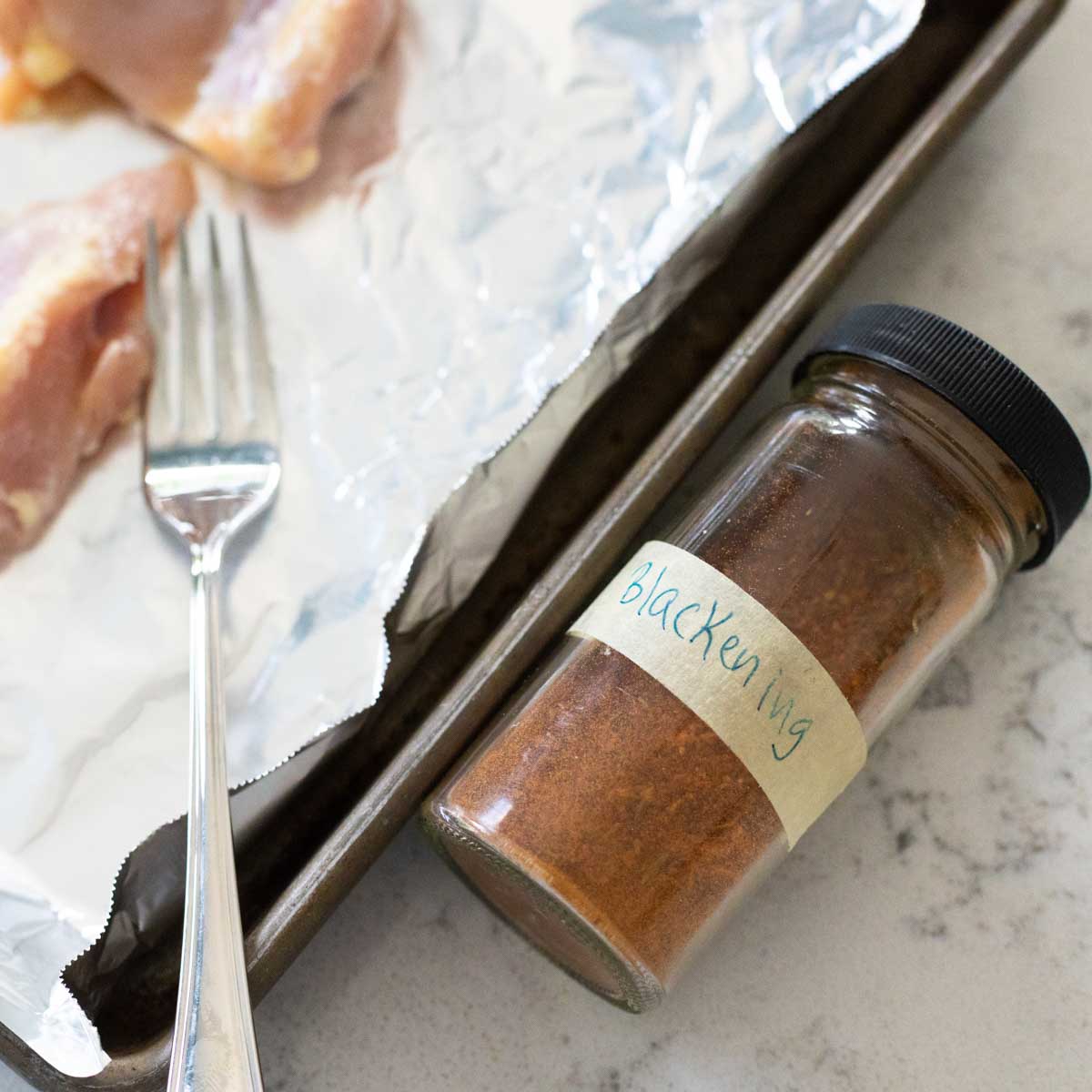 A jar of homemade blackening seasoning lays on the counter next to a baking pan with chicken.