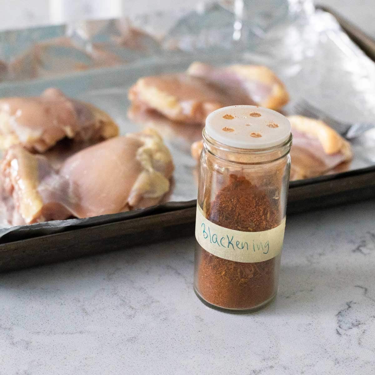 The spice jar sits next to the chicken thighs on a roasting pan.