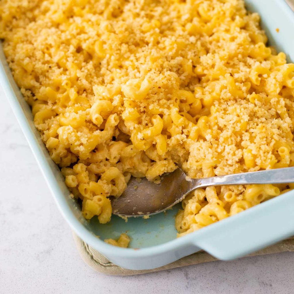 A blue baking dish filled with mac and cheese with bread crumb topping. A serving spoon has removed a portion to show the creamy elbow noodles.