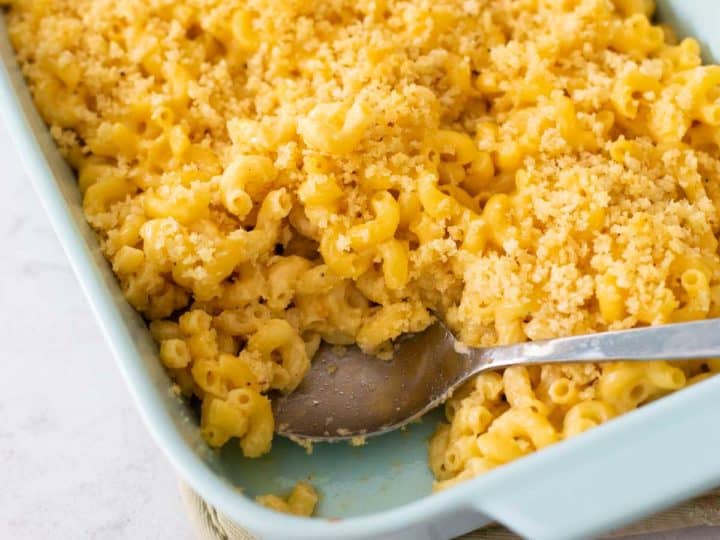 A blue baking dish filled with mac and cheese with bread crumb topping. A serving spoon has removed a portion to show the creamy elbow noodles.