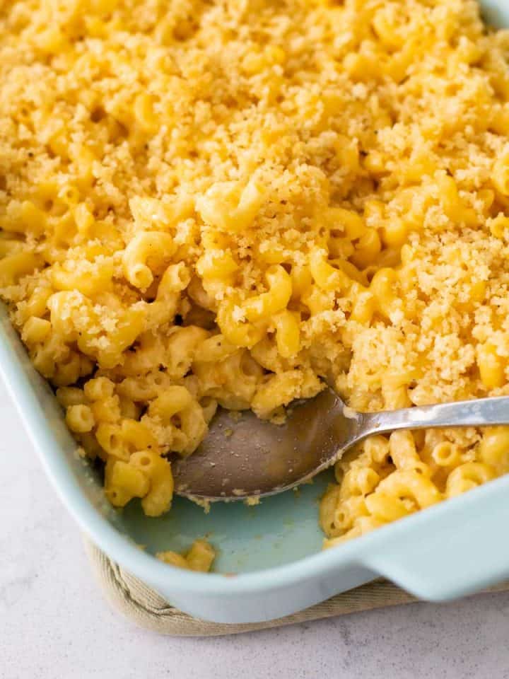 A blue baking dish filled with mac and cheese with bread crumb topping. A serving spoon has removed a portion to show the creamy elbow noodles.