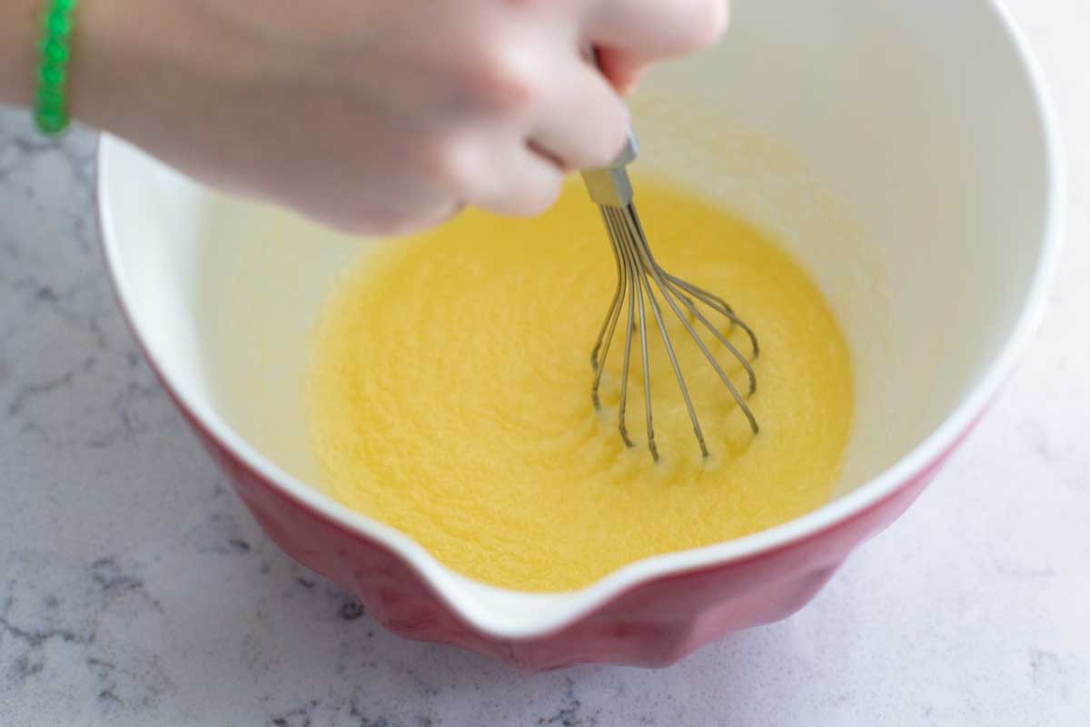 A girl's hand is whisking melted butter and sugar together in a batter bowl.