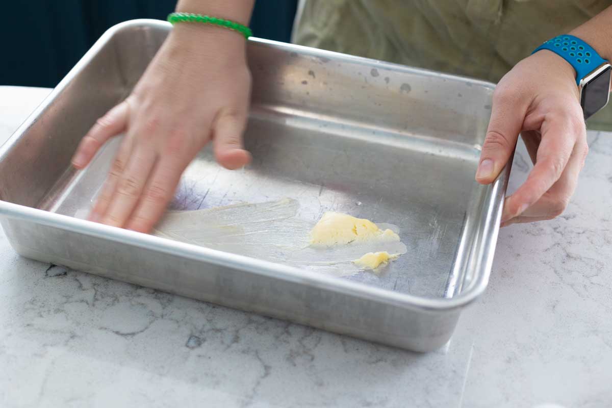A girl's hands are rubbing softened butter inside a metal baking pan.