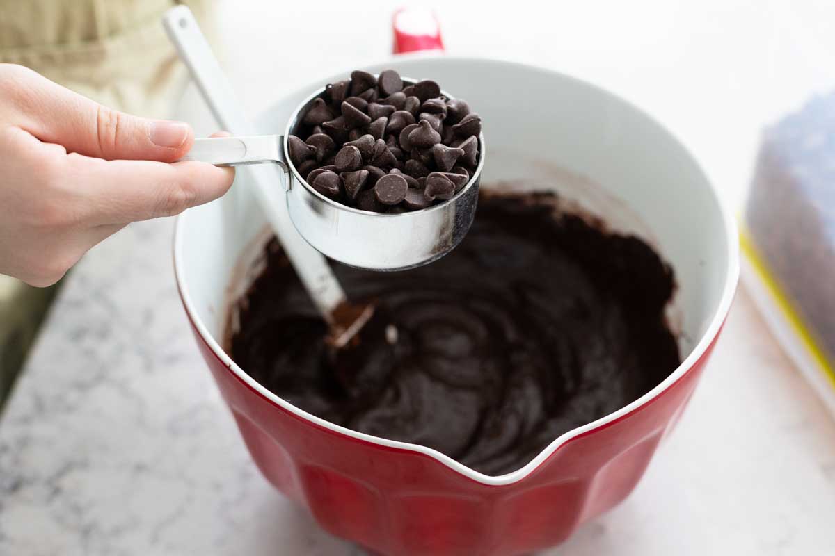 A girl's hand is about to pour a measuring cup of chocolate chips into the brownie batter.