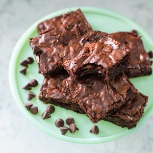 A round platter of a pile of chocolate brownies with a bite taken out of the one on top and a sprinkle of chocolate chips to the side.